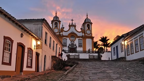 As igrejas barrocas e ruas de pedra de cidades históricas mineiras evocam o cenário romântico e clássico da série "Bridgerton". Igreja barroca amarela e branca ao entardecer com rua de paralelepípedos e casarões coloniais.