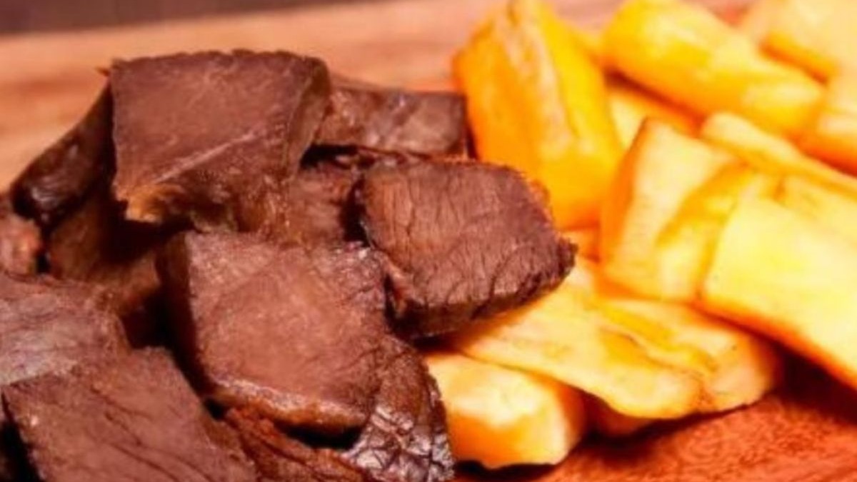 Peda&ccedil;os de carne de sol escura ao lado de macaxeira frita amarela em prato de madeira.