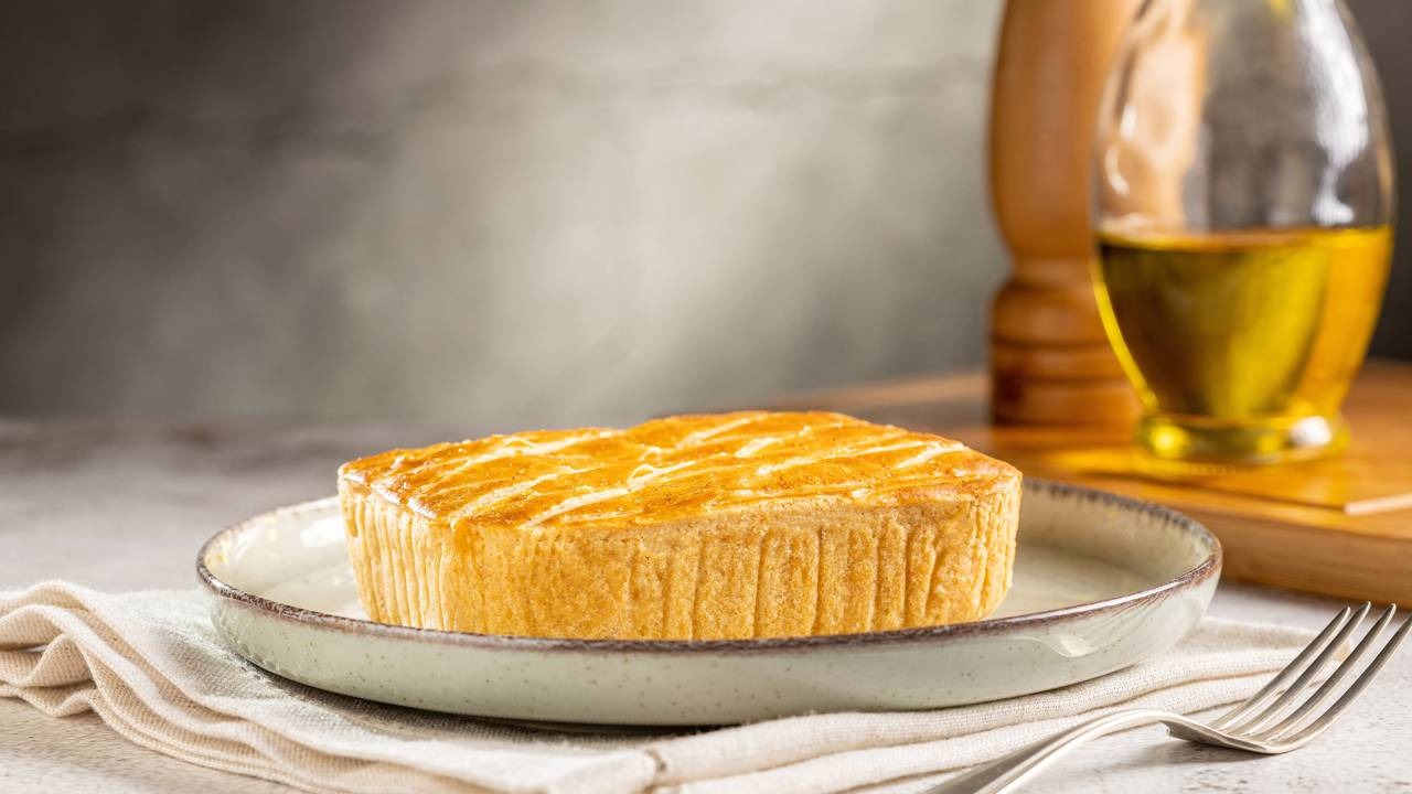 O empadão de frango, com sua massa dourada e recheio cremoso, é a estrela da receita. Empadão de frango retangular com massa dourada em prato, talher e azeite ao fundo