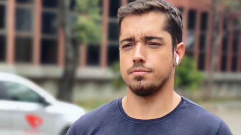 Caíque Verli, o repórter da TV Gazeta que faleceu nesta quinta, em registro que remete à sua dedicação profissional. Homem de camiseta azul, com barba e fone de ouvido branco na orelha direita