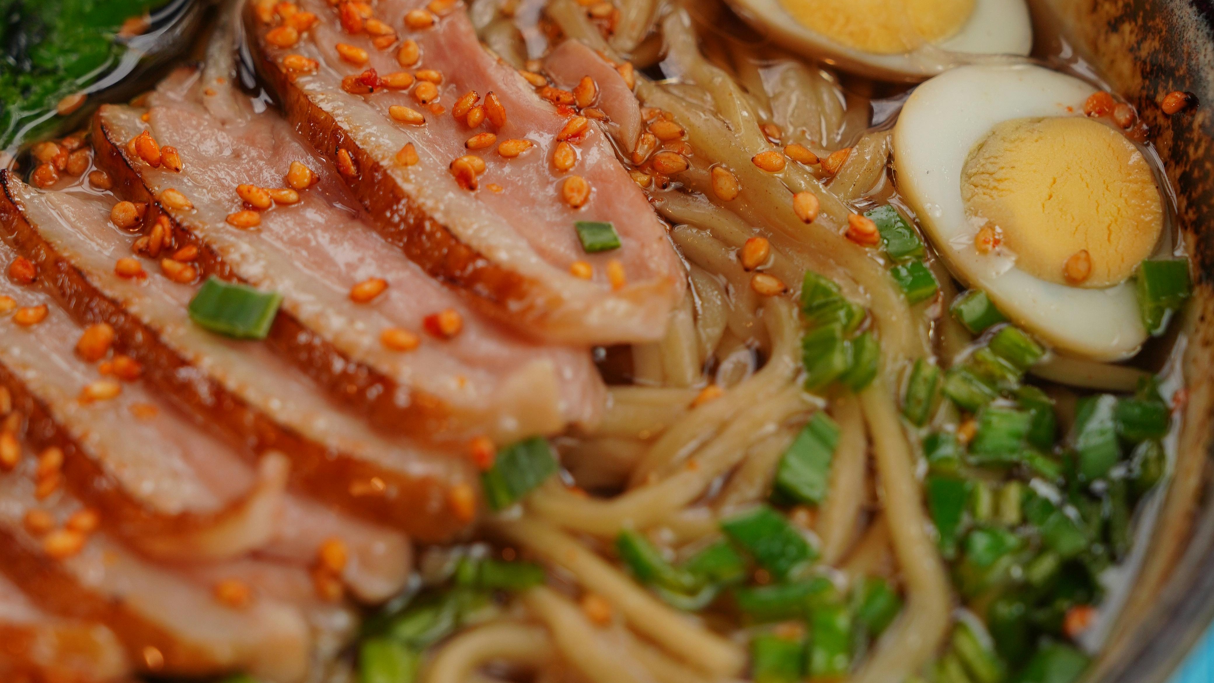Close-up de tigela de rámen com macarrão, fatias de carne, ovo cozido, cebolinha e sementes.