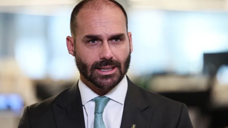 Close-up de Eduardo Bolsonaro, com barba, terno escuro e gravata azul, falando seriamente