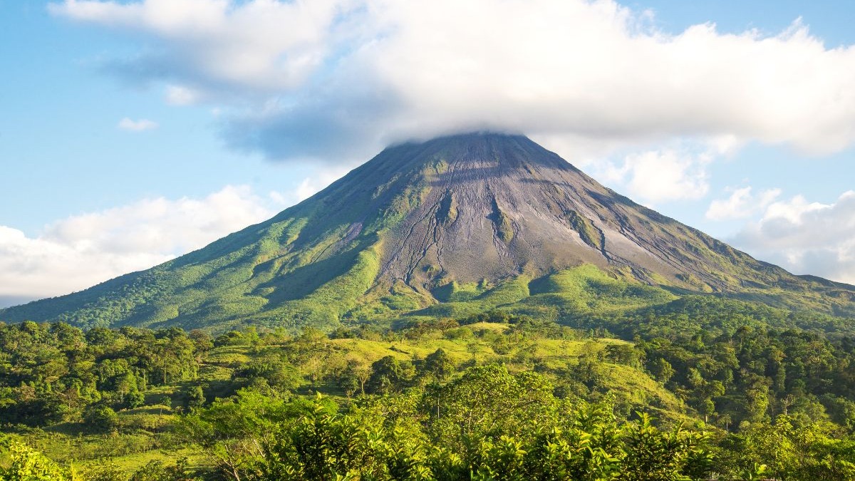 A exuberância natural do vulcão Arenal, na Costa Rica, destaca-se como polo ecoturístico. Vulcão cônico Arenal com nuvens no cume, encostas verdes e densa floresta tropical.