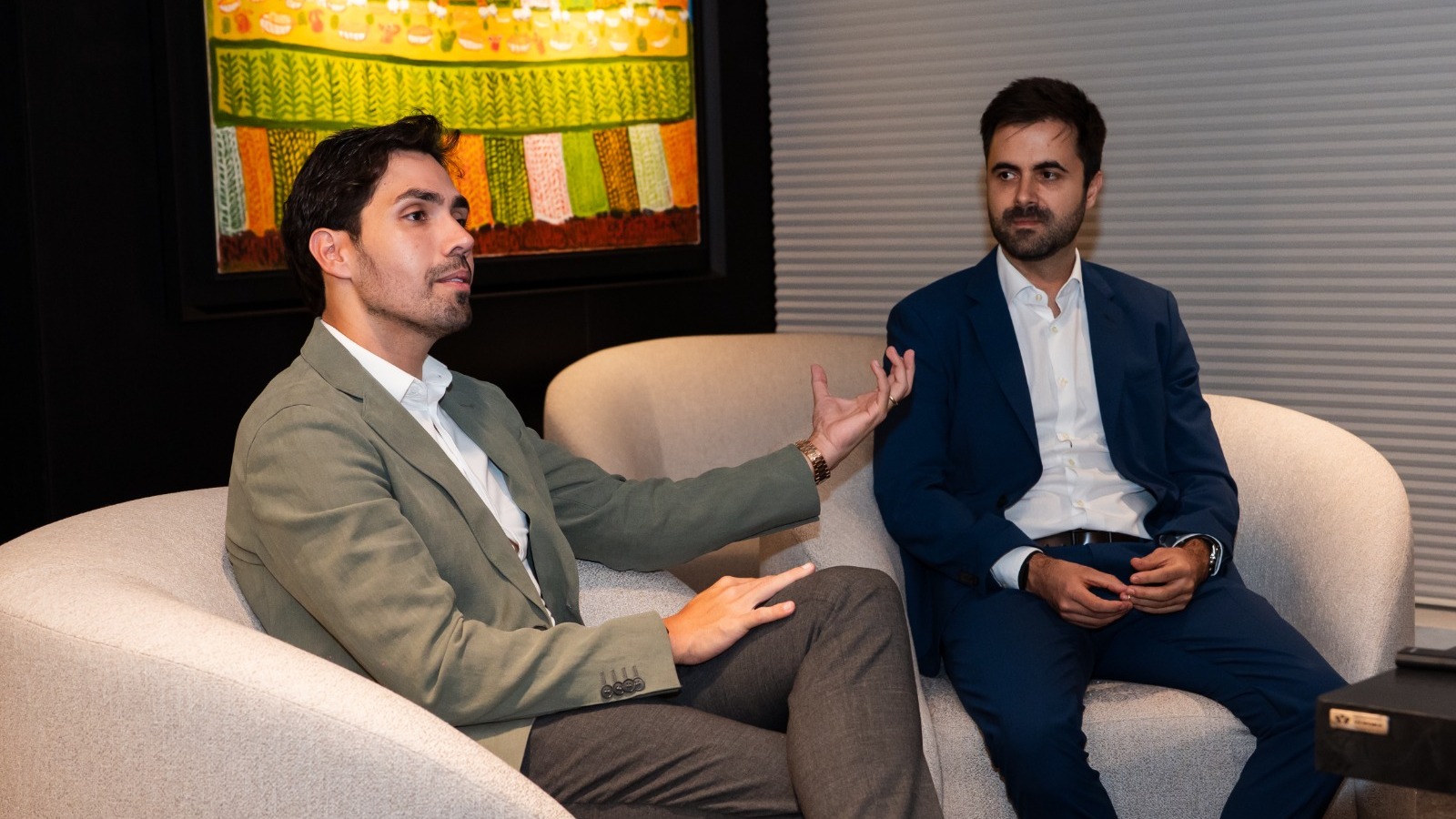 Dois homens sentados em poltronas claras conversam, um gesticula com a m&atilde;o direita