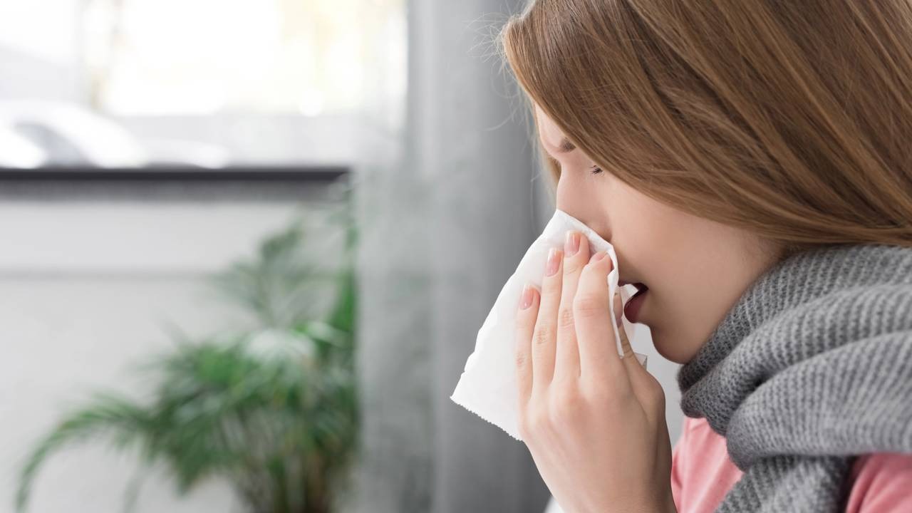Sintomas respiratórios como a rinite podem piorar com a frente fria, um dos efeitos da mudança climática no organismo. Mulher de perfil assoando o nariz com lenço de papel branco, usando cachecol cinza.