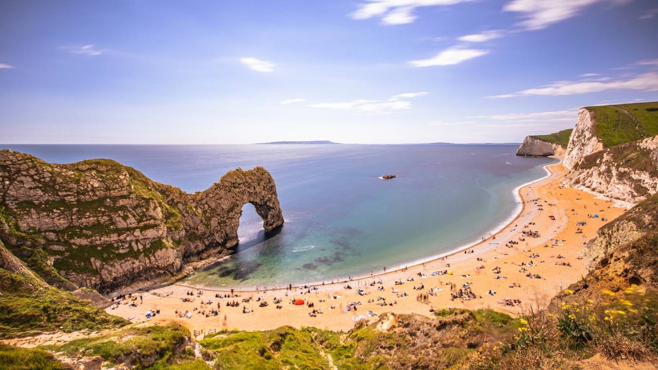 A icônica Durdle Door, na Inglaterra, parte da Jurassic Coast, um dos locais onde a erosão revela fósseis e segredos da história geológica. Vista aérea panorâmica de uma praia de areia, mar azul-turquesa, falésias rochosas e arco natural