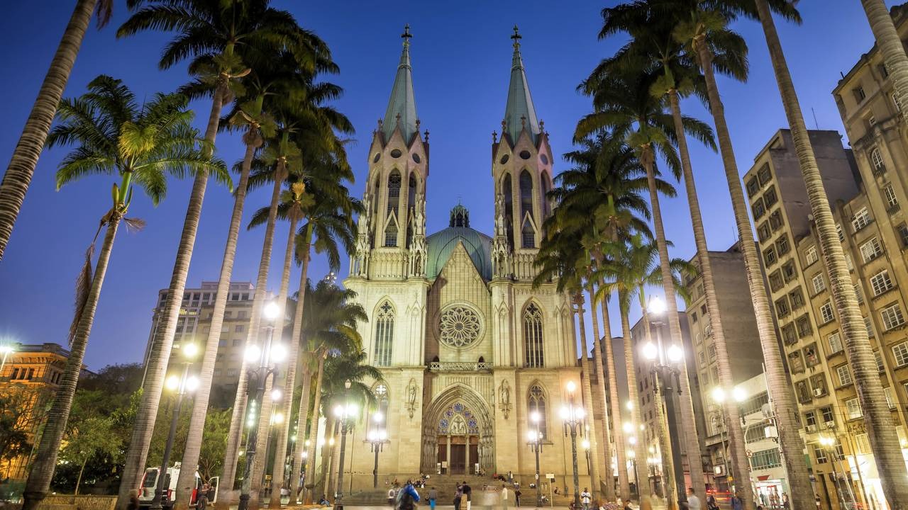 A Catedral da Sé, um dos cartões-postais de São Paulo, marca o início do roteiro histórico do segundo dia na capital. Catedral da Sé gótica iluminada à noite, palmeiras e postes de luz.