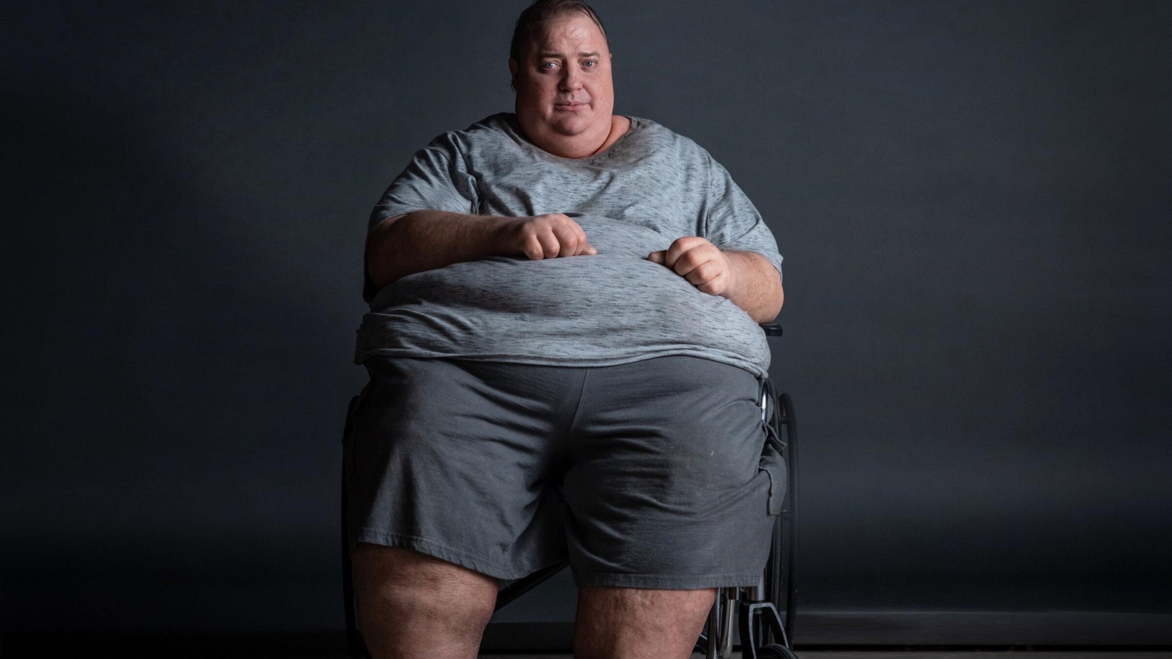 Brendan Fraser, caracterizado com próteses que simulam obesidade severa, está sentado em uma cadeira de rodas, olhando para a frente. Ele veste uma camiseta cinza e shorts cinzas. O fundo é liso e escuro.
