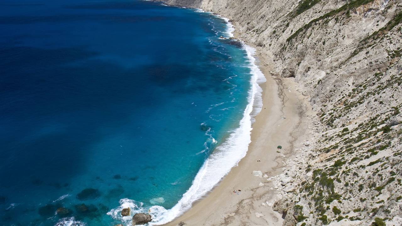 A grandiosidade das paisagens das Ilhas Jônicas, como a famosa Praia de Myrtos, conecta o viajante à saga de Ulisses Créditos: depositphotos.com / Panos_Karas Vista aérea de praia grega com mar azul-turquesa, areia clara e falésias rochosas íngremes com vegetação