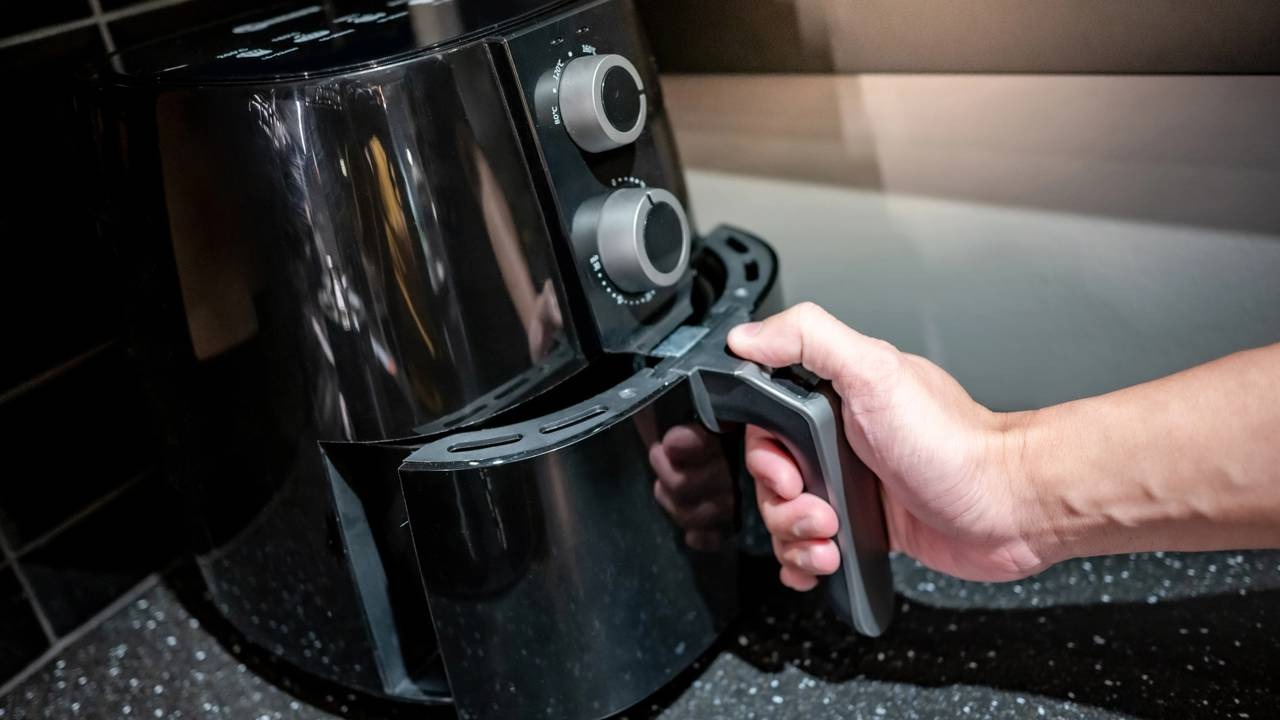Verificar a airfryer é essencial para garantir o cozimento homogêneo dos alimentos, conforme detalhado no teste de circulação de ar. Uma pessoa puxa o cesto de uma airfryer preta com botões prateados sobre uma bancada escura