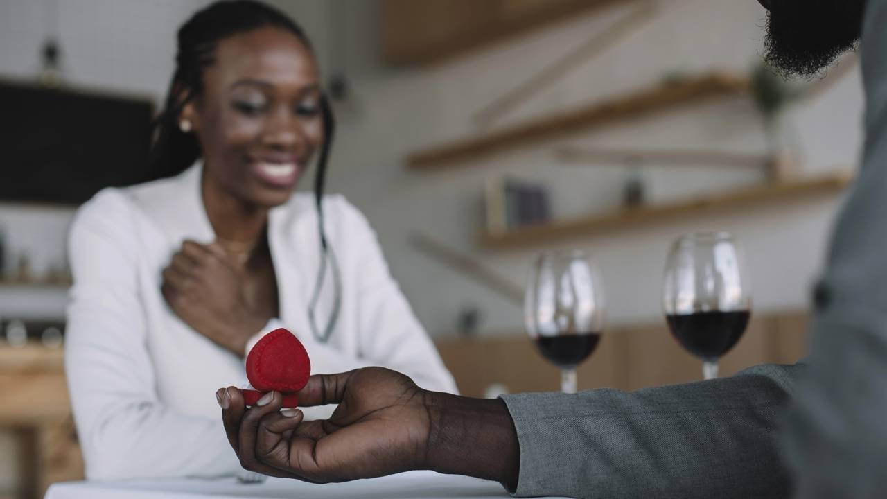 Um gesto romântico de amor, como um pedido de casamento, simboliza a busca pelo presente perfeito no Dia dos Namorados. Mão de homem negro oferece anel de noivado em caixa vermelha a mulher negra feliz.