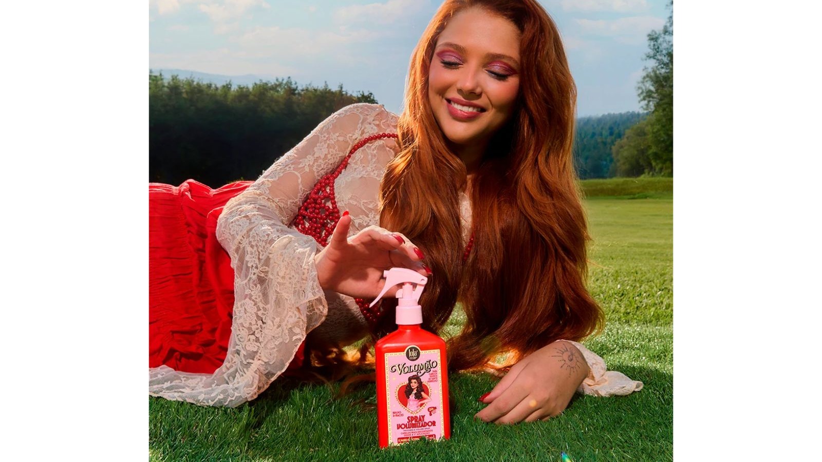 Mulher ruiva sorridente deitada na grama segura um frasco vermelho do Lola Volum&atilde;o Spray Volumizador