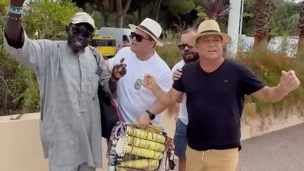 O cantor Leonardo interage com um vendedor ambulante em Cannes, na França, cantando e dançando durante viagem. Quatro homens sorrindo, um vendedor com mercadorias e Leonardo de chapéu em Cannes