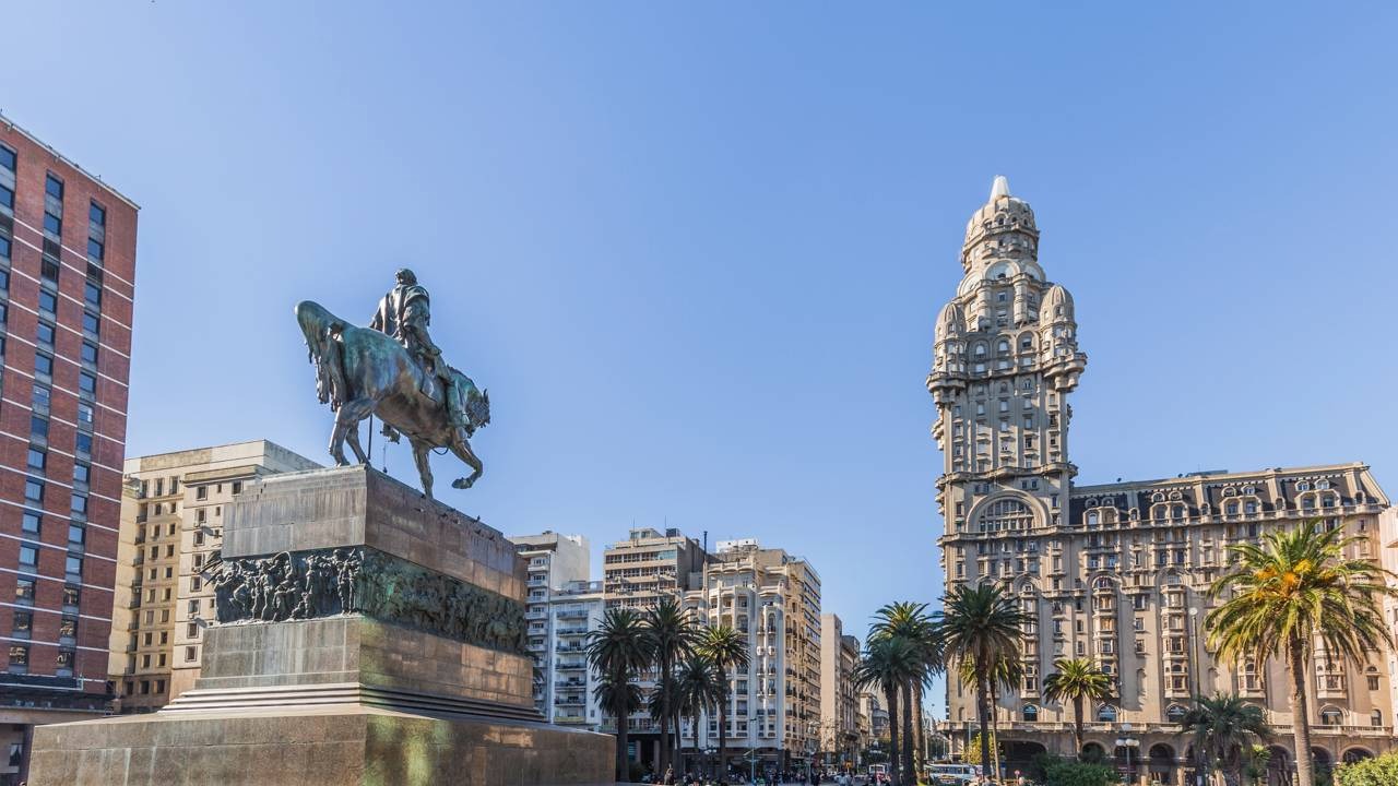 Montevidéu, capital do Uruguai, país apontado como o melhor destino para aposentados na pesquisa da Global Citizen Solutions. Estátua equestre de bronze em praça, prédio histórico ornamentado, palmeiras e edifícios urbanos