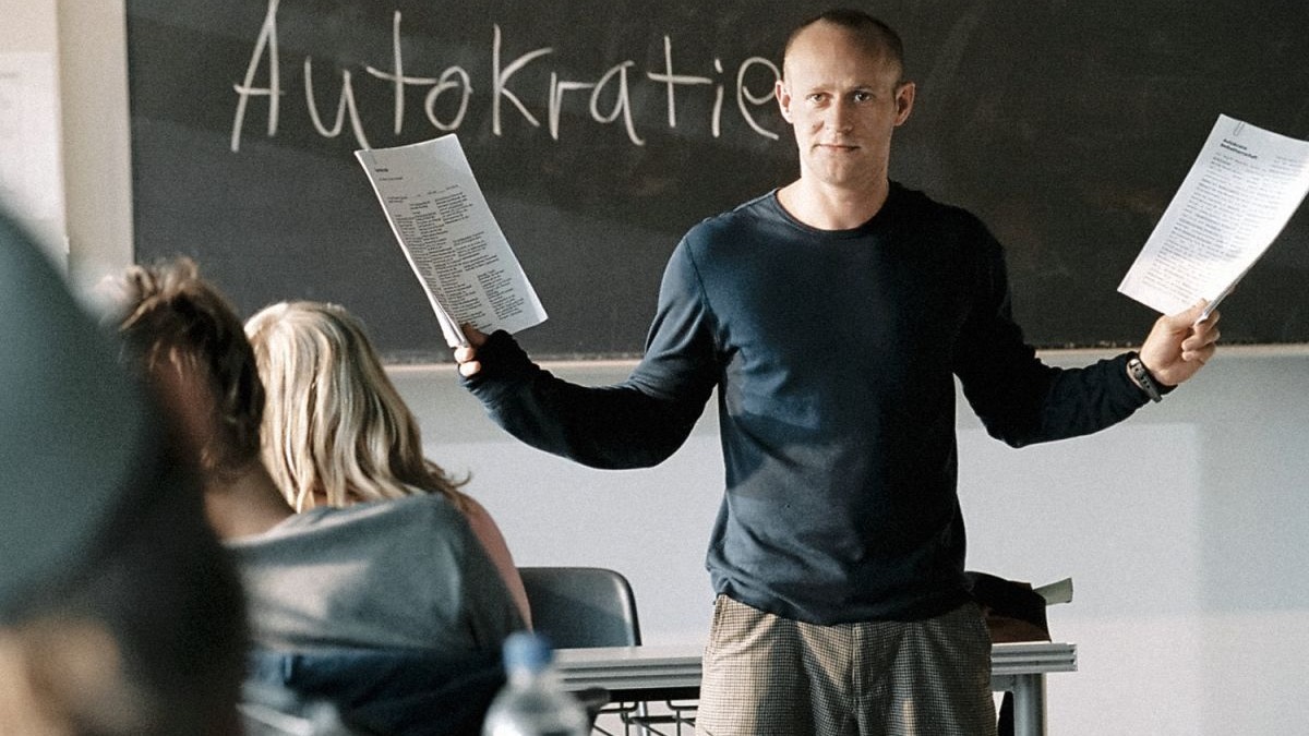 Professor calvo em sala de aula, segurando pap&eacute;is, com 