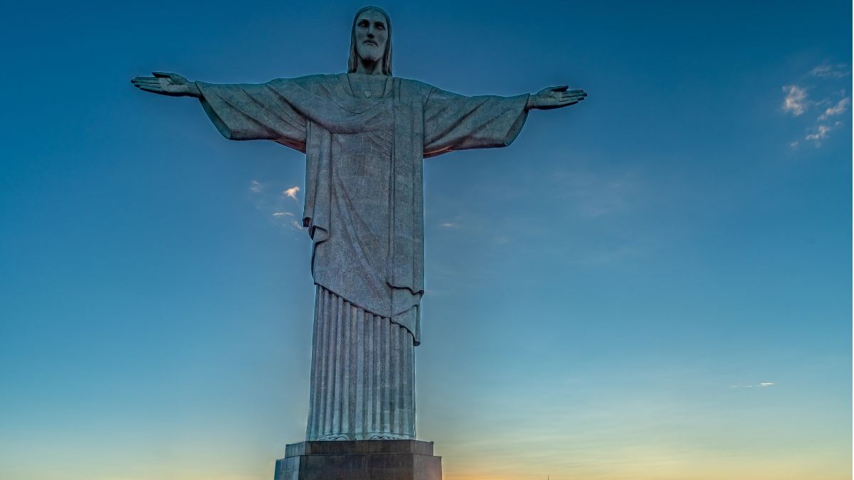 O Cristo Redentor, símbolo do Rio de Janeiro e da fé brasileira, integra a lista das Sete Maravilhas do Mundo Moderno. Estátua do Cristo Redentor em pedra, braços abertos, contra céu azul claro.