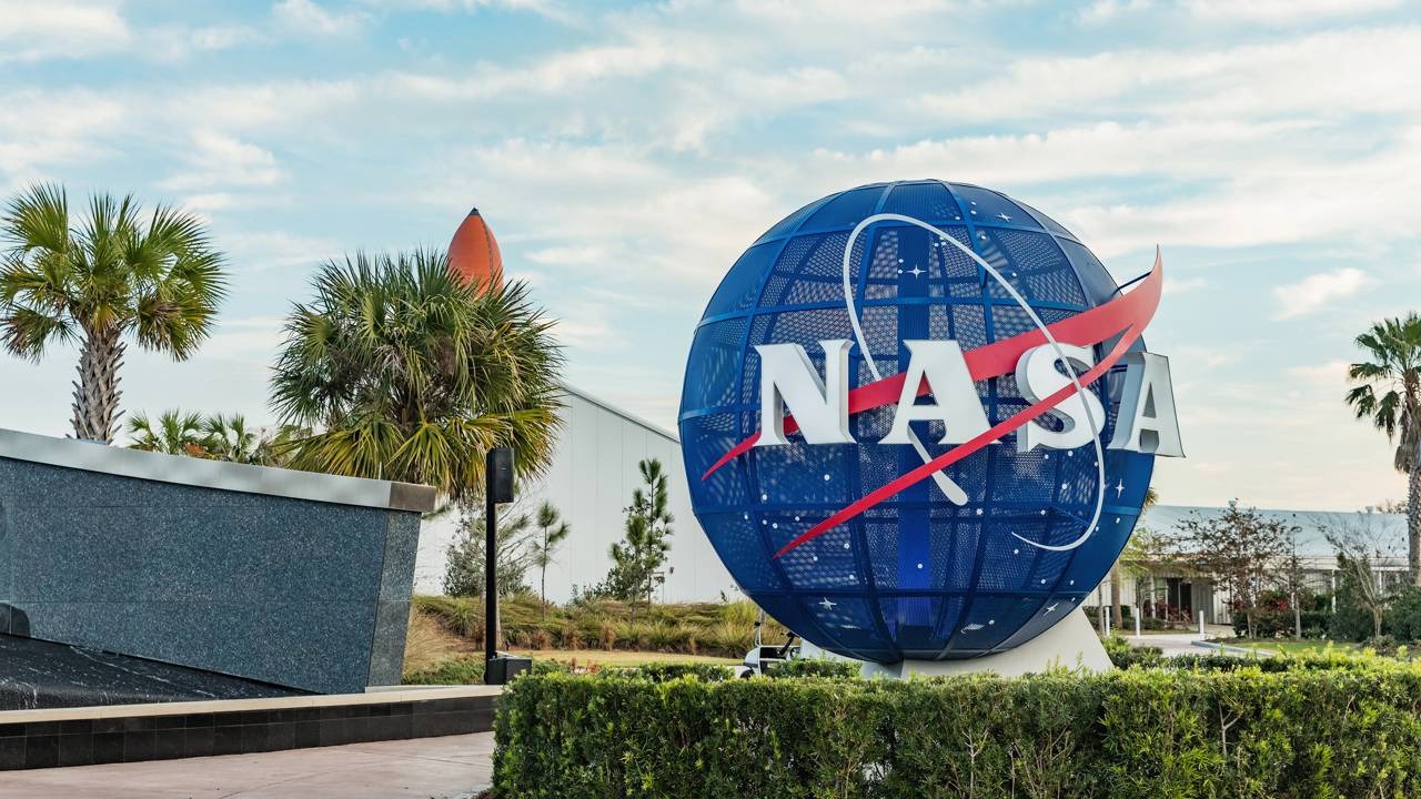 A NASA lidera pesquisas sobre asteroides como Bennu, desvendando componentes essenciais para a vida no universo. Globo azul com logo da NASA em branco e vermelho, palmeiras e arbustos verdes, sob céu azul com nuvens