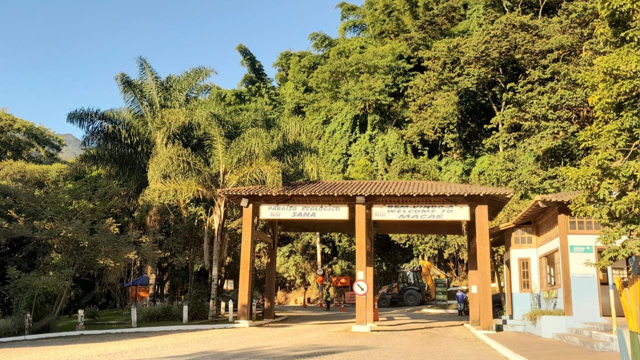 O portal de entrada para o Paraíso Ecológico Sana dá as boas-vindas aos visitantes do refúgio de ecoturismo em Macaé. Portão de madeira com placas 'Paraíso Ecológico Sana' e 'Welcome to Macaé', cercado por mata.