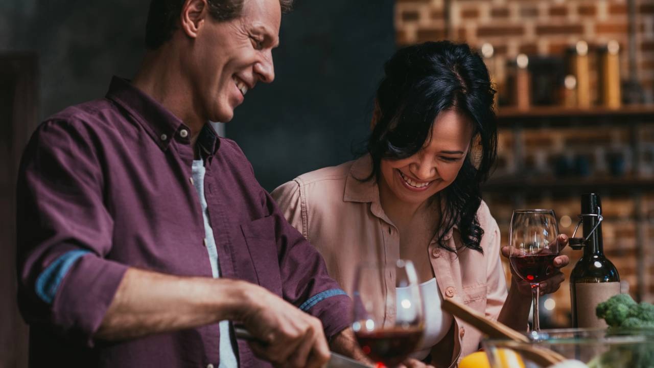 Casal se diverte preparando a refeição com vinho, um cenário ideal para um risoto romântico e descomplicado no Dia dos Namorados. Homem e mulher sorrindo, preparando comida com vinho tinto em uma cozinha.