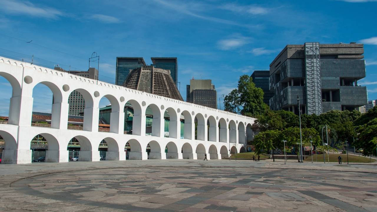 Os Arcos da Lapa, um dos maiores símbolos do Rio de Janeiro, representam o coração boêmio onde a paixão pelo futebol toma conta. Arcos brancos da Lapa no Rio de Janeiro sob céu azul com prédios e árvores.