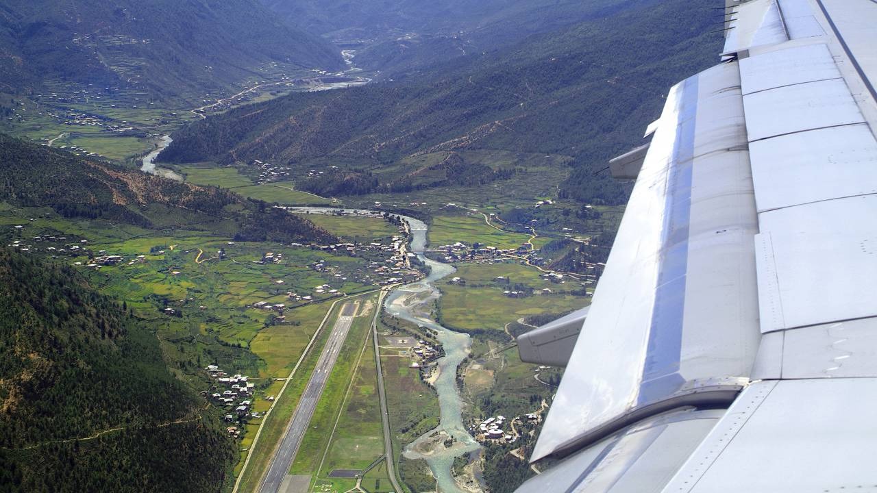 A vista da asa de um avião durante a complexa aproximação ao Aeroporto de Paro, no Butão, um dos mais desafiadores do mundo. Asa de avião sobre vale montanhoso com rio sinuoso, pista de aeroporto e pequenas construções.