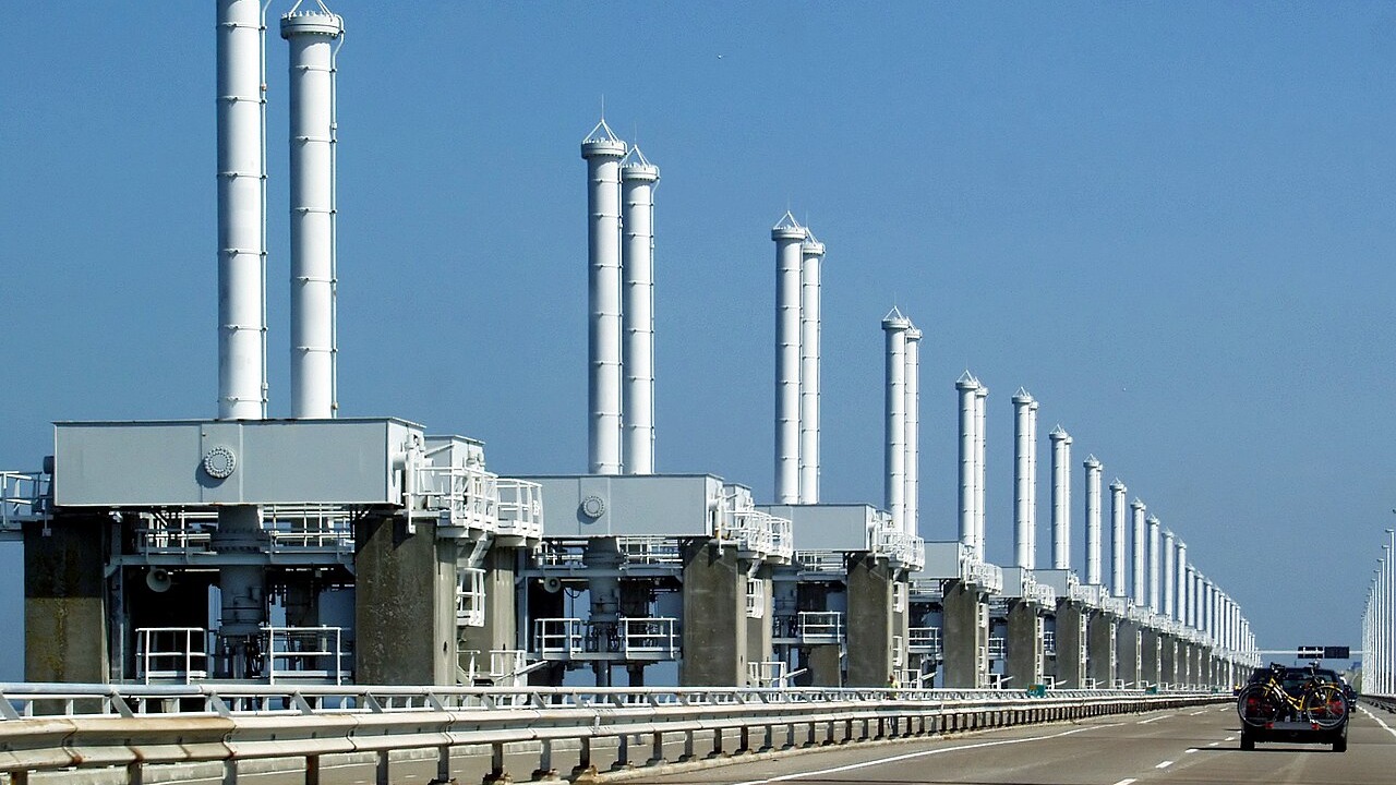 A Oosterscheldekering, parte do Plano Delta holandês, exemplifica a engenharia que protege o país contra inundações. Carro em autoestrada ladeada por extensa barreira contra inundações com pilares brancos sob céu azul.