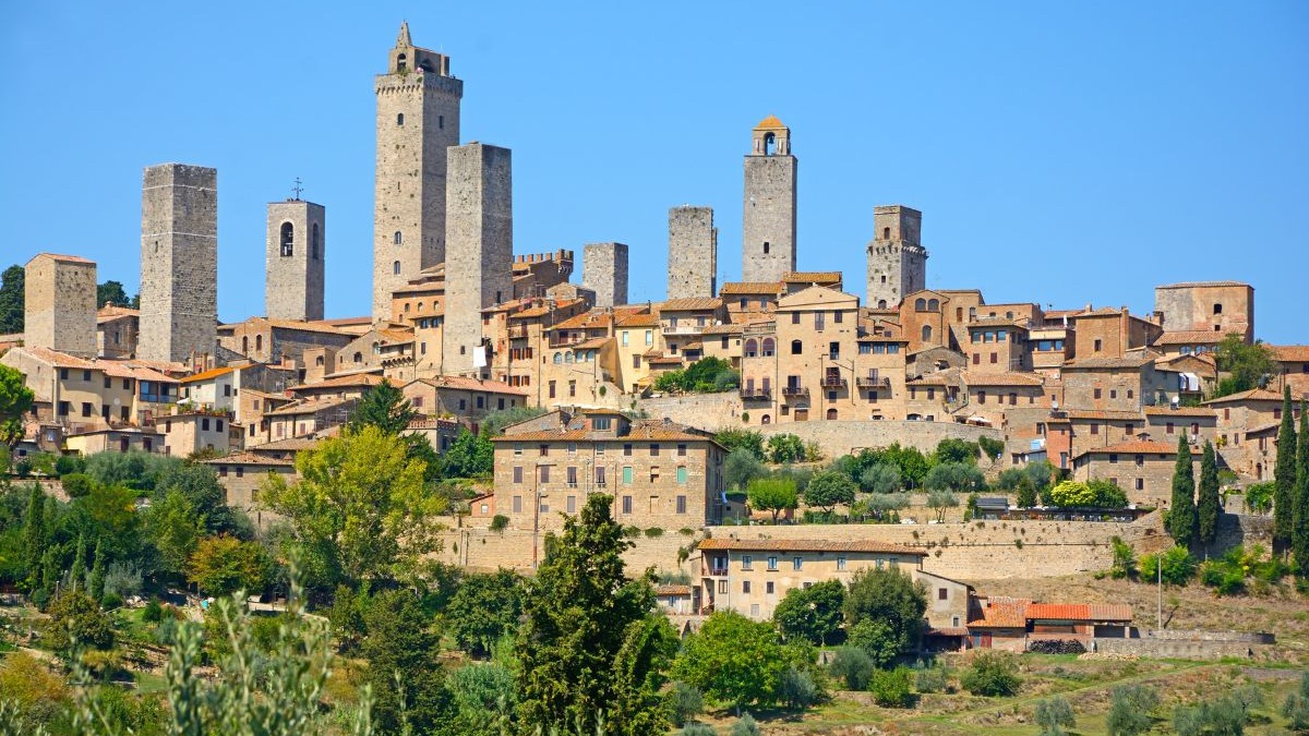 As imponentes torres medievais de San Gimignano, na Toscana, destacam-se no cenário histórico do vilarejo italiano. Vista panorâmica de San Gimignano com várias torres medievais e casas de pedra sob céu azul.
