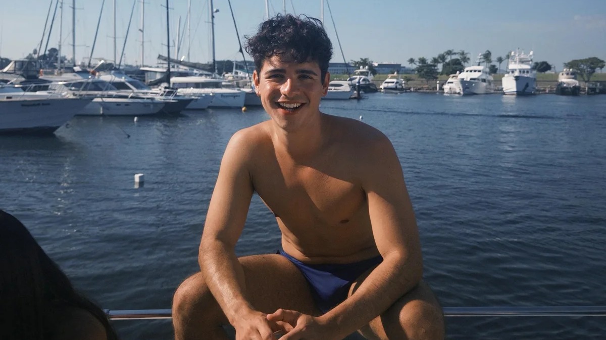 O ator João Victor Gonçalves, que gerou polêmica ao falar sobre o Rio, em cenário que remete aos locais que elogiou na cidade Jovem sorridente, sem camisa e de sunga azul, sentado em barco com marina cheia de embarcações
