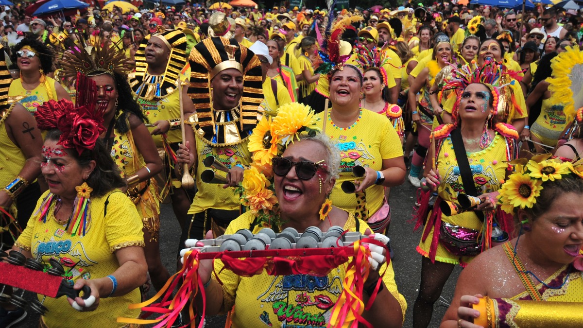 Foliões sorridentes em fantasias amarelas com maquiagem e instrumentos musicais no carnaval de rua.
