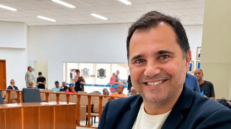 Jefferson Dias da Silva, secretário de Desenvolvimento Econômico de Queimados, falecido após ser baleado em abordagem policial - Foto; Reprodução/Redes Sociais Homem sorridente de blazer azul e camisa clara em ambiente interno com pessoas ao fundo