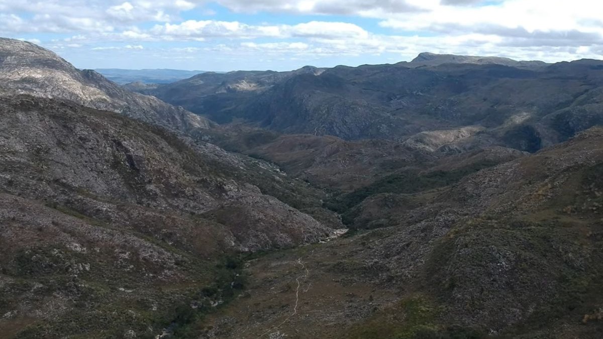 Vista a&eacute;rea de um vale sinuoso entre montanhas rochosas e vegeta&ccedil;&atilde;o esparsa sob c&eacute;u nublado.