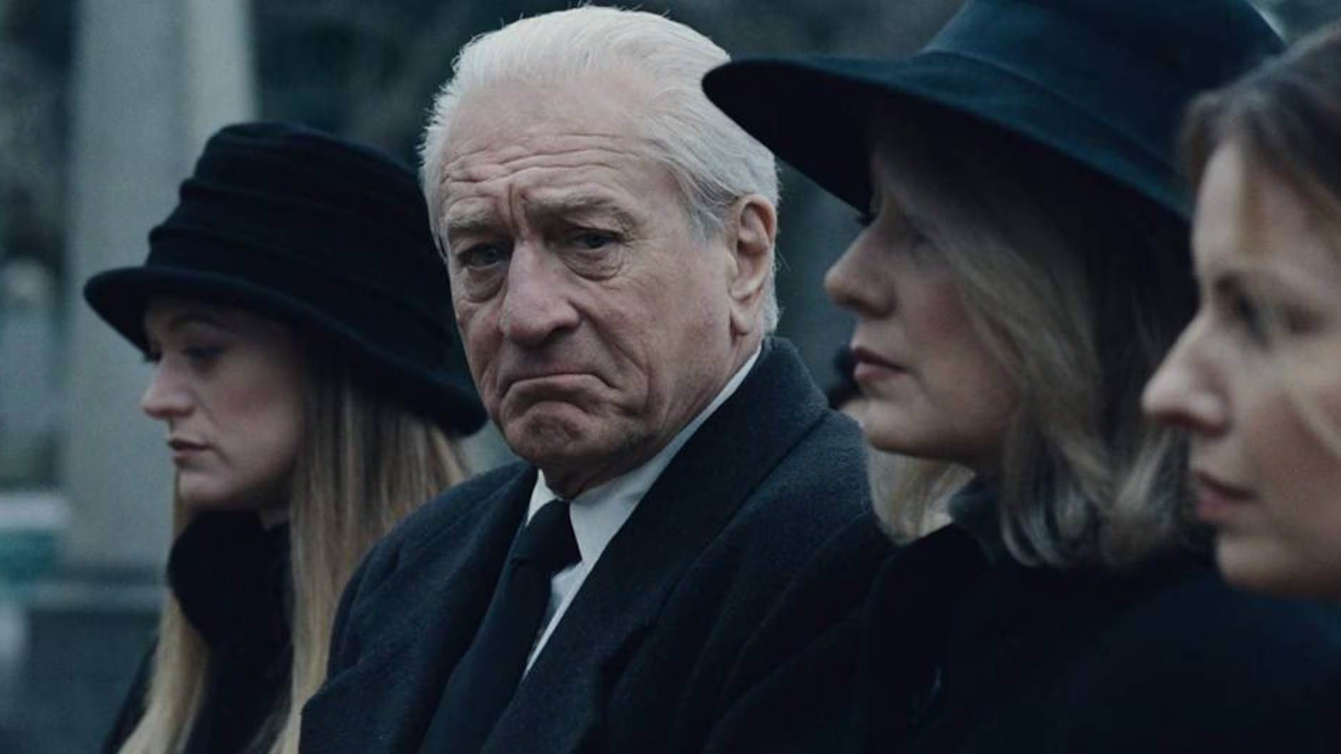 Robert De Niro em cena de 'O Irlandês', filme que o reuniu novamente com Martin Scorsese para um drama reflexivo sobre a máfia. Foto: Divulgação Robert De Niro com cabelos brancos e expressão sombria em terno escuro, entre mulheres de chapéu.