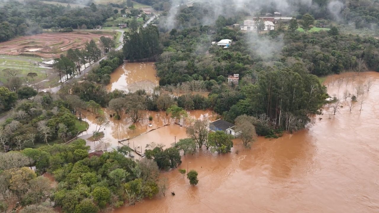 Área do Rio Grande do Sul severamente impactada por enchentes, com casas e vegetação submersas. Vista aérea de área rural inundada com casas, árvores e estrada submersas por água barrenta