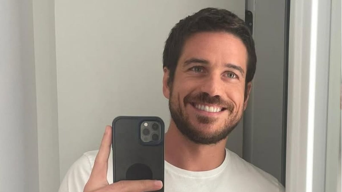 Marco Pigossi, ator escalado para a nova série do Prime Video "Obsessão" e "Clube Hedonê", em momento de descontração. Foto: Reprodução Homem sorridente com barba, camiseta branca, segura celular em espelho