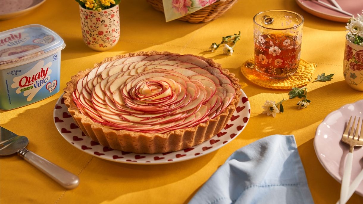 Torta de ma&ccedil;&atilde; florida em prato decorado, Qualy Vita, ch&aacute;, flores e talheres em mesa amarela.