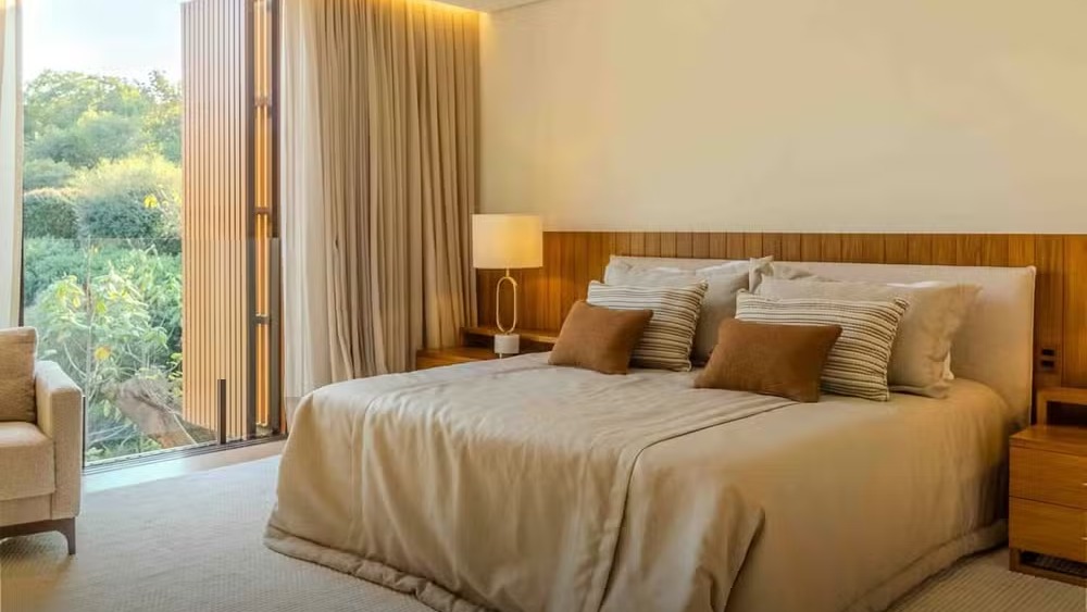 Cama com roupa de cama clara, cabeceira de madeira, luminária e grande janela para árvores