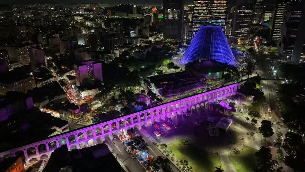 Arcos da Lapa à noite, um dos palcos da Semana de Arte do Rio com shows e atividades gratuitas. Vista aérea noturna dos Arcos da Lapa iluminados em roxo e Catedral Metropolitana em azul, Rio de Janeiro