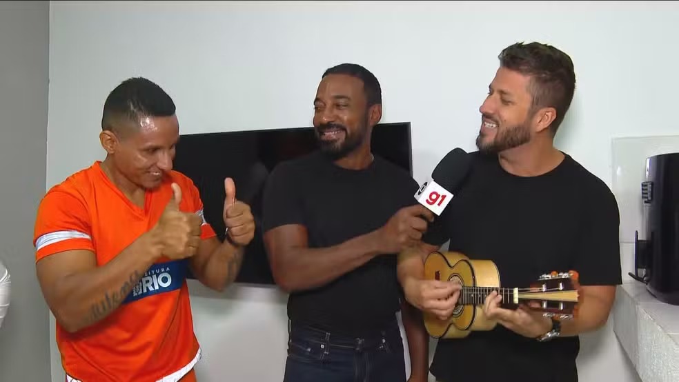 O músico Felipe Branco homenageia o gari Fabiano Cândido da Silva com uma canção após a devolução dos pertences. Três homens sorrindo. Um gari, um músico com cavaquinho e microfone, e um terceiro homem