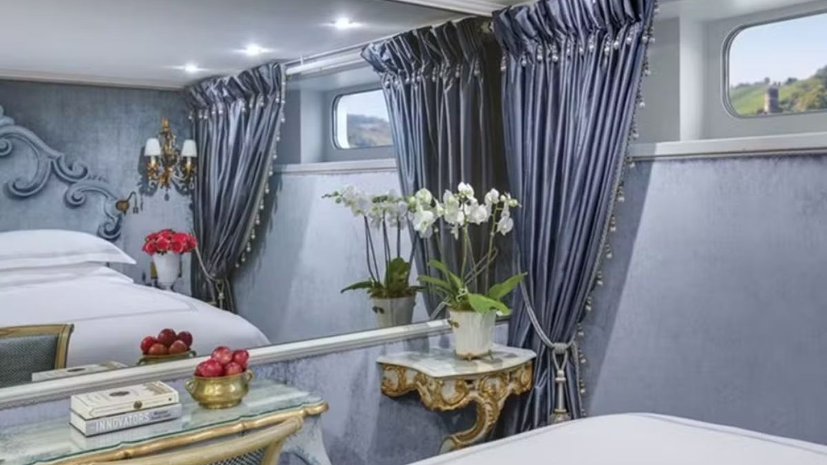 Su&iacute;te de cruzeiro luxuosa com cama, cortinas azuis, orqu&iacute;deas e janelas com vista.