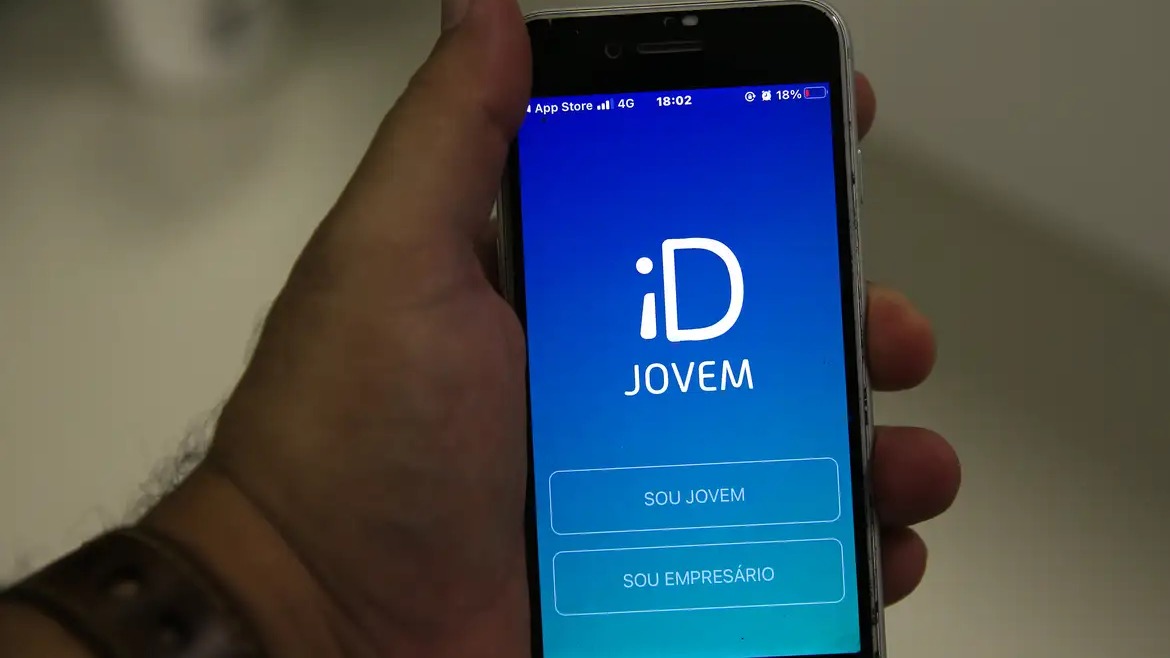 O aplicativo iD Jovem, um dos benefícios do CadÚnico, oferece vantagens para jovens de 15 a 29 anos. Mão segura celular com app iD Jovem, botões 'SOU JOVEM' e 'SOU EMPRESÁRIO' visíveis.
