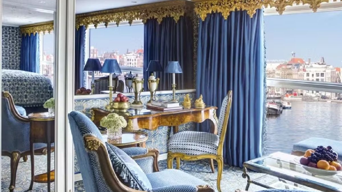 Su&iacute;te de navio luxuosa azul e dourada, cama, m&oacute;veis cl&aacute;ssicos, vista para rio e cidade.