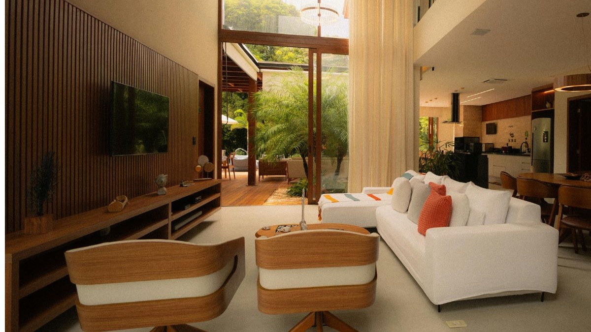 Sala de estar moderna com sof&aacute; branco, painel ripado de madeira e janelas para jardim.