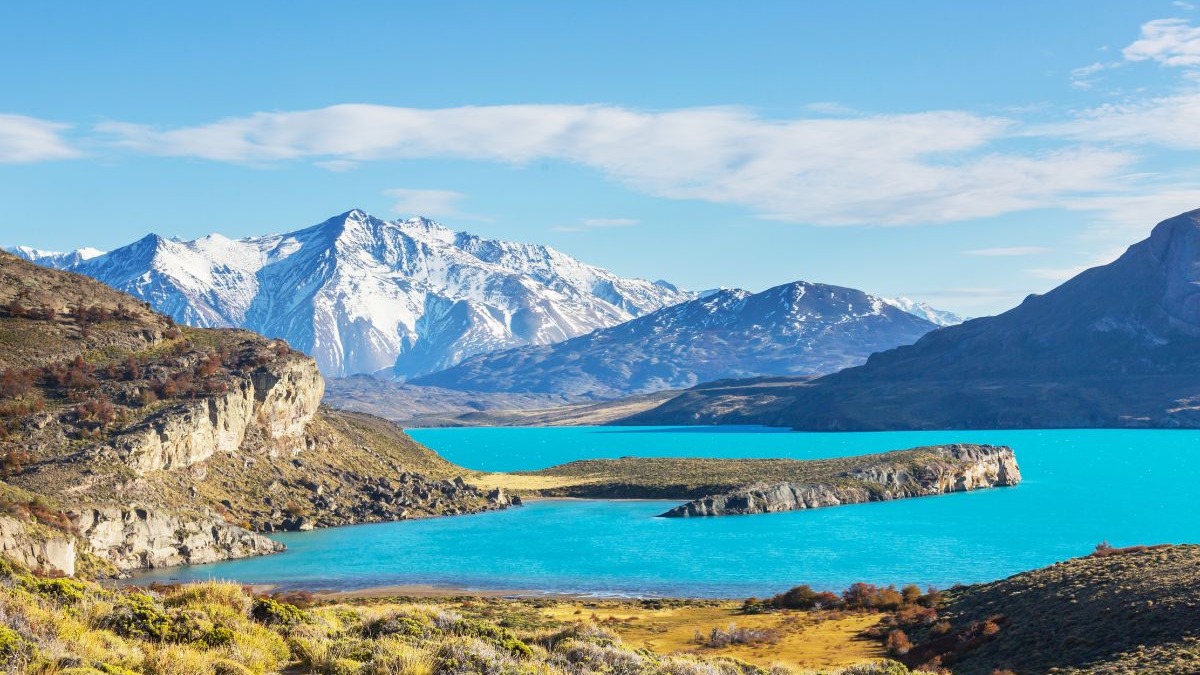 Lago azul-turquesa entre montanhas rochosas e nevadas sob céu azul claro.