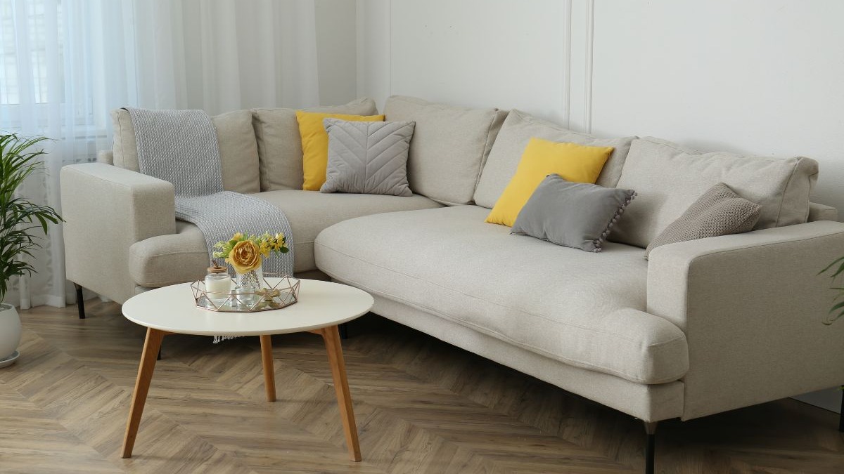 Sof&aacute; secional bege, almofadas amarelas e cinzas, mesa de centro, planta e cortinas leves.