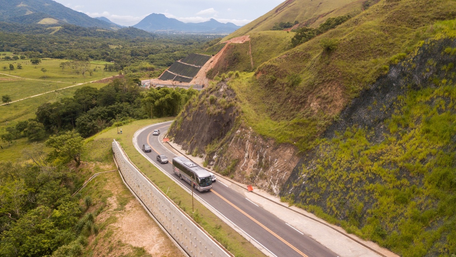Usuários do Bilhete Único Intermunicipal podem usufruir do benefício em linhas de ônibus que conectam diferentes municípios do estado. Ônibus e carros em estrada montanhosa com vegetação luxuriante e encostas verdes.