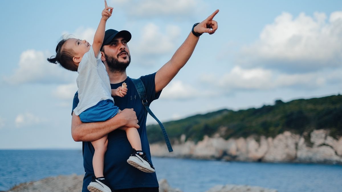 A relação entre pais e filhos, com a escolha de nomes homônimos, pode carregar um caminho de expectativas Pai com boné segurando filho pequeno, ambos apontando para cima, com mar e montanhas