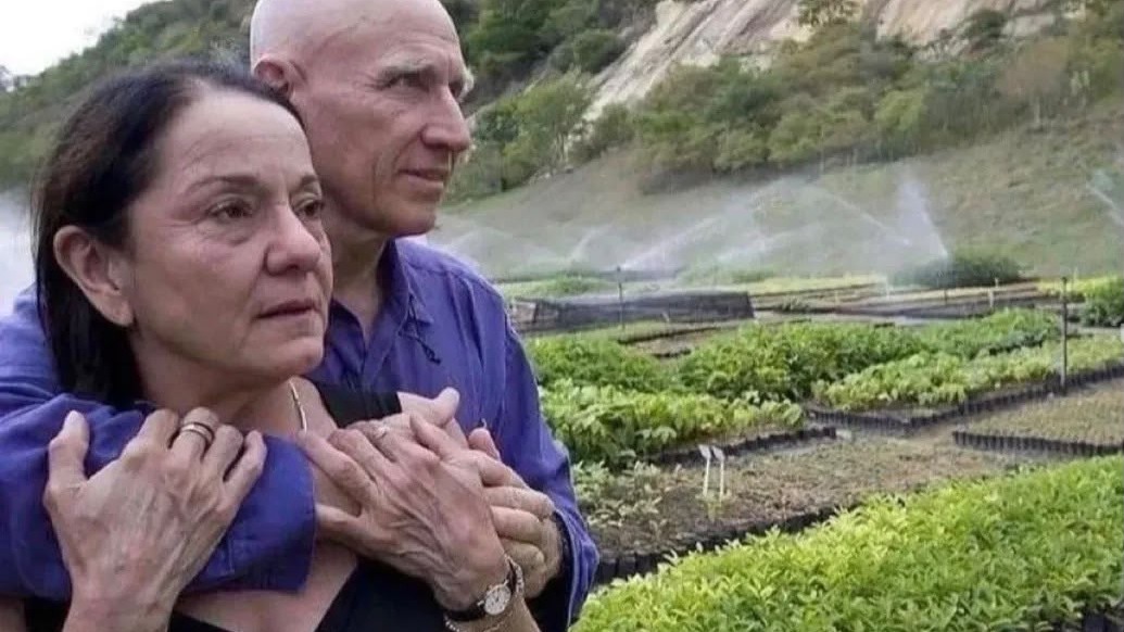Lélia e Sebastião Salgado no viveiro de mudas, o ponto de partida do projeto que transformou a fazenda em floresta exuberante Um homem careca abraça uma mulher de cabelo escuro em um viveiro de plantas com irrigação