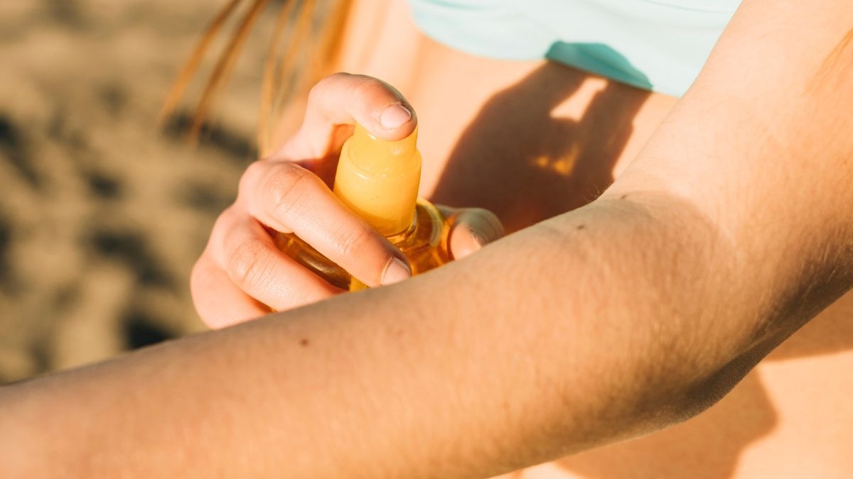 M&atilde;o aplicando spray de frasco amarelo no bra&ccedil;o, sob luz solar forte.
