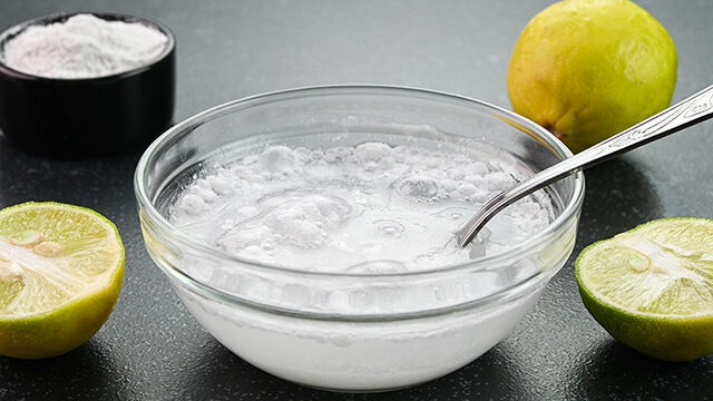 A solução de limão e bicarbonato é um dos métodos caseiros eficazes para tentar recuperar roupas com manchas recentes de água sanitária. Tigela de vidro com mistura efervescente de bicarbonato de sódio e limão, com uma colher.