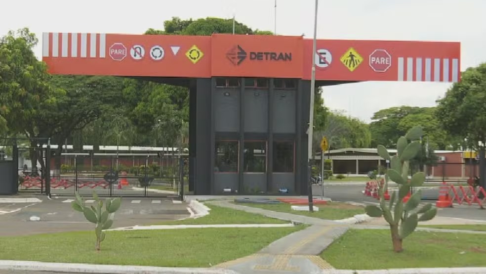 A sede do Detran-GO, que foi condenado a ressarcir motoristas por taxas indevidas, é vista na imagem Entrada do Detran com portaria, letreiro Detran, placas de trânsito, gramado, cactos e árvores
